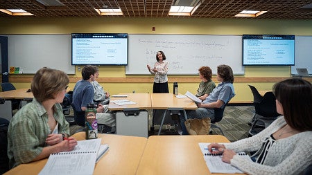 French professor in front of class with white boards and screens and students in foreground